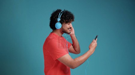Photo for Cheerful handsome young latin or arabian man in casual watching videos on smartphone, using new headset, looking at cell phone and smiling, in coral t-shirt on blue studio background, copy space - Royalty Free Image