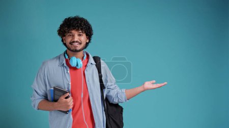 Téléchargez les photos : Jeune étudiant indien ou espagnol homme avec sac à dos sur fond bleu studio pointant vers le côté pointant vers le côté présentant votre produit. - en image libre de droit