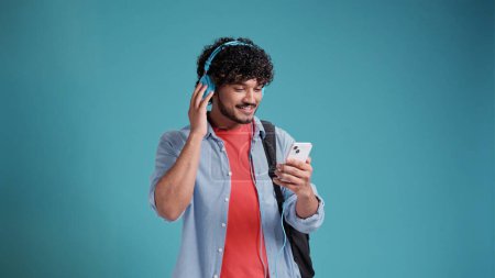 Téléchargez les photos : Jeune étudiant indien ou espagnol écoutant de la musique avec écouteurs et smartphone sur fond de studio bleu. - en image libre de droit
