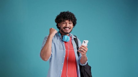Foto de Joven estudiante indio o español con una mochila se regocija en el éxito y la victoria con un teléfono inteligente en sus manos. En un fondo de estudio azul - Imagen libre de derechos