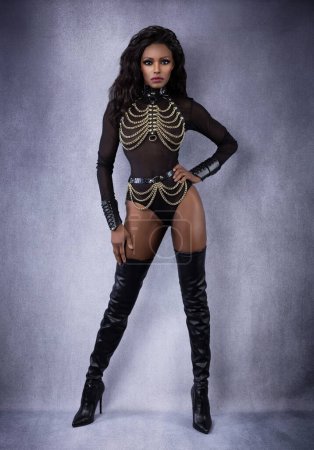 Beautiful sexy black dominant woman in leather chain harness outfit and thigh high boots posing full length in studio on gray mottled background