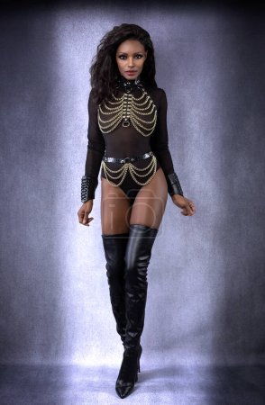 Beautiful sexy black dominant woman in leather chain harness outfit and thigh high boots posing full length in studio on gray mottled background