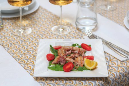 Téléchargez les photos : Assiette carrée avec mini calamars et garniture de feuilles vertes et tomates cerises servies avec un couteau et une fourchette avec un verre d'eau et de vin - en image libre de droit