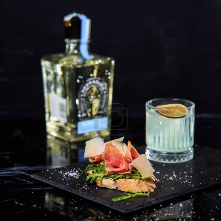 Photo for Tequila, cocktail and bruschetta with prosciutto on a black ceramic board on a black marble table - Royalty Free Image