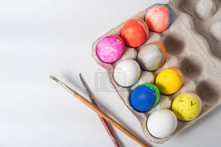 Photo for Colorful painted Easter eggs on a cardboard tray with paint brushes on a white background. Happy Easter concept. - Royalty Free Image