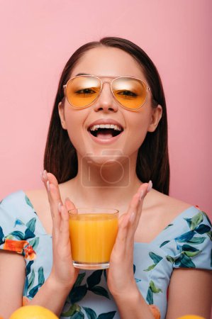 Téléchargez les photos : Femme montrant du verre de jus d'orange et souriant à la caméra seule. Jeune beau modèle caucasien isolé sur fond rose - en image libre de droit