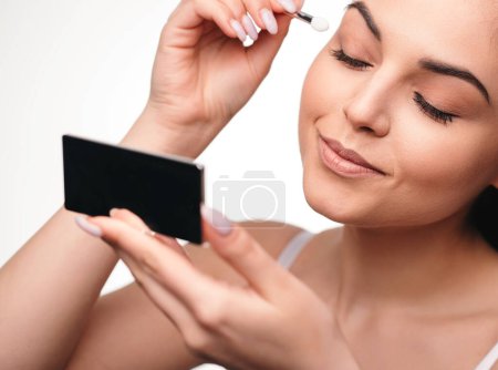 Foto de Chica feliz mirando espejo sosteniendo cepillo mientras se aplica maquillaje. Hermosa chica morena bonita aplicando maquillaje sobre fondo blanco - Imagen libre de derechos
