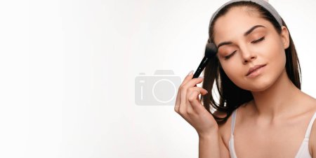 Téléchargez les photos : Jolie jeune femme utilisant un pinceau cosmétique pour appliquer un maquillage décoratif sur son visage, fond de studio blanc. Millennial femme mettre sur blusher - en image libre de droit