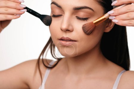 Téléchargez les photos : Portrait de beauté de jeune femme brune caucasienne appliquant des cosmétiques avec pinceau de maquillage isolé sur fond blanc - en image libre de droit