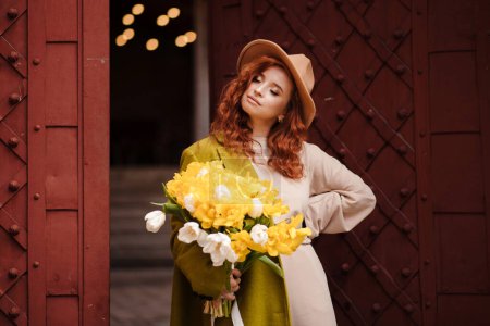 Foto de Mujer atractiva joven en sombrero, con los ojos cerrados posando cerca del edificio viejo con puertas rojas antiguas. En las manos mujer sosteniendo un ramo de tulipanes amarillos. Moda y estilo de vida. - Imagen libre de derechos