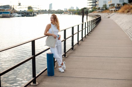 Téléchargez les photos : Jeune femme avec de longs cheveux blonds debout au bord de la rivière et regardant loin après l'entraînement de yoga. Concept du sport et des personnes - en image libre de droit