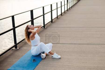 Foto de Cuerpo completo de hembra relajada sonriente sentada en una esterilla de yoga en un puente cerca del terraplén del río y descansando después del entrenamiento. - Imagen libre de derechos