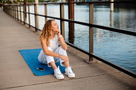 Téléchargez les photos : Femme détendue souriante assise sur un tapis de yoga sur un pont près d'un remblai de rivière et regardant l'eau pendant qu'elle se repose après l'entraînement - en image libre de droit