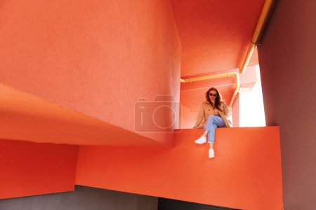 Téléchargez les photos : Hipster fille posant contre le mur rugueux de la rue rouge. Femme en lunettes de soleil dans le style vestimentaire urbain assis dans la rue - en image libre de droit