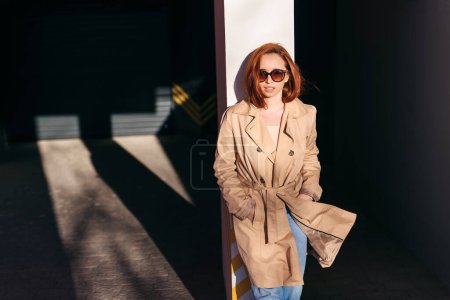 Téléchargez les photos : Femme aux cheveux roux élégant posant à l'arrière-plan urbain. Ginger girl en lunettes de soleil dans le style de vêtements urbains debout dans la rue - en image libre de droit