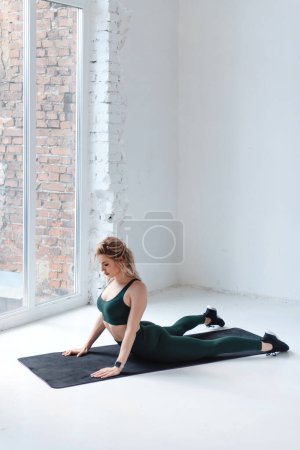 Photo for Young blonde woman in sportswear practicing yoga in a white hall opposite large window. Fashion sports fitness slender girl performs exercises. Workout on the mat - Royalty Free Image