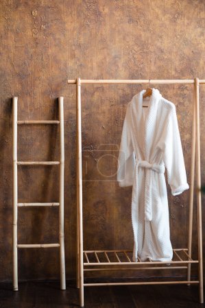 Photo for Close up of small wooden ladder standing near floor hanger with white soft bathrobe. Stylish details in modern interior. - Royalty Free Image