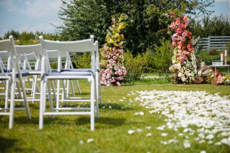 Téléchargez les photos : Cérémonie de mariage en plein air avec arche en fleur colorée au milieu et chaises en bois blanc debout sur les côtés. Belle zone pour marié et mariée sur la nature. - en image libre de droit