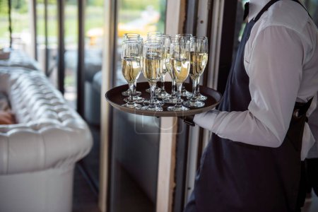Photo for Close up of male waiter in uniform and black gloves carrying tray with glasses full of champagne. Restaurant worker serving alcoholic drinks for guests at wedding celebration. - Royalty Free Image