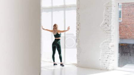 Foto de Vista trasera de la joven deportista en top verde, polainas deportivas de pie en un estudio blanco y mirando a la ventana. Chica fitness con el pelo rubio - Imagen libre de derechos