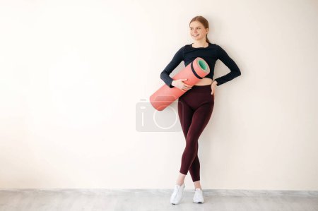 Foto de Longitud completa de la mujer fitness sonriente de pie en un estudio de fitness que lleva una esterilla de yoga. Retrato de una joven en un gimnasio. - Imagen libre de derechos
