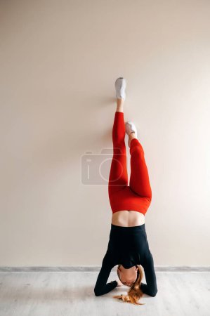 Foto de Vista frontal de una joven rubia haciendo un soporte de mano sobre una pared blanca con espacio para copiar. Slim mujer deportiva en rojo tigth pantalones leggings practicar equilibrio pose - Imagen libre de derechos