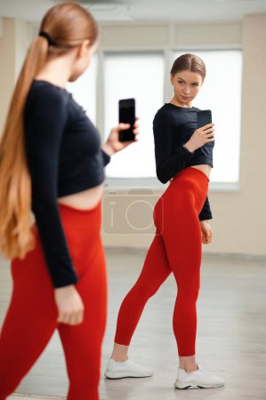 Téléchargez les photos : Sport, fitness, mode de vie, technologie et concept de personnes - jeune femme en legging rouge serré avec smartphone prenant miroir selfie dans la salle de gym - en image libre de droit