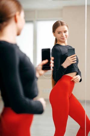 Téléchargez les photos : Fit femme bronzée en rouge et noir vêtements de sport, corps parfait, abdos motivation, à la maison prendre photo selfie sur le téléphone dans le miroir pour les médias sociaux, histoires, vertical - en image libre de droit