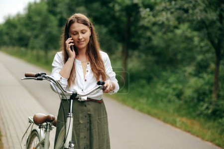 Photo for Attractive young lady taking short break after riding bicycle in forest and talking on smartphone. Charming woman with long brown hair leading healthy lifestyle and using ecological way of transport. - Royalty Free Image