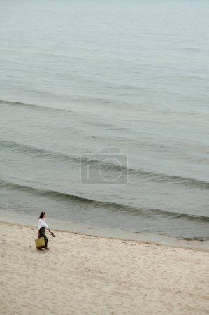 Téléchargez les photos : Vue ci-dessus de la femme élégante marchant sur la plage de sable le long du littoral en saison froide et regardant sur les vagues. Dame aux cheveux foncés en vêtements décontractés portant des chaussures et un sac de paille dans les mains. - en image libre de droit
