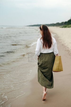Foto de Vista trasera de una hembra morena paseando por la costa de arena con una gran bolsa de playa en la mano. Mujer vestida casualmente mirando en las olas y disfrutando de caminar en la isla vacía. Concepto de armonía e inspiración. - Imagen libre de derechos