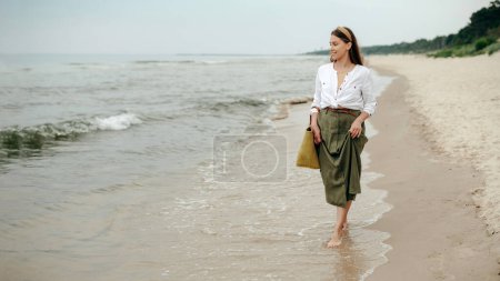 Photo for Young attractive woman dressed in olive color skirt and white shirt walking barefoot along seashore. Smiling lady in headband enjoying warm waves on deserted sandy beach. - Royalty Free Image