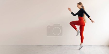 Foto de Vista lateral de la atleta haciendo ejercicios de fitness y saltando alto en el centro de aeróbic. Dama activa llena de energía usando top ajustado con leggins rojos y preparándose para competiciones deportivas. - Imagen libre de derechos