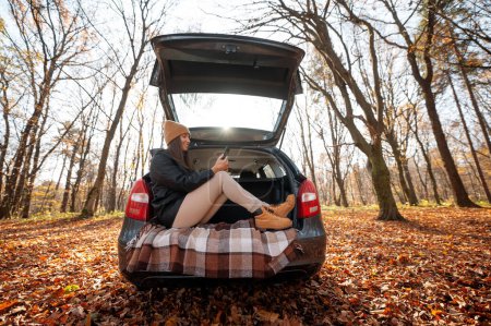 Foto de Mujer feliz disfrutando con teléfono inteligente en el maletero del coche durante la mañana de otoño. Bosque de otoño viaje en coche concepto - Imagen libre de derechos