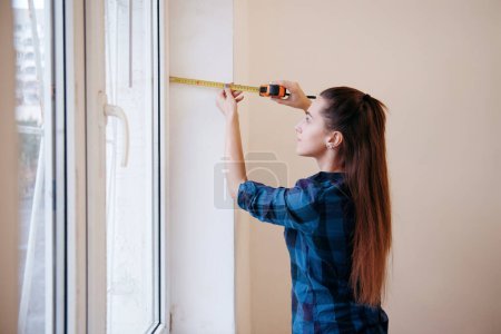 Téléchargez les photos : Jeune femme brune mesurant le rebord de fenêtre avec un ruban à mesurer à la maison. Rénovation. Amélioration de la maison - en image libre de droit