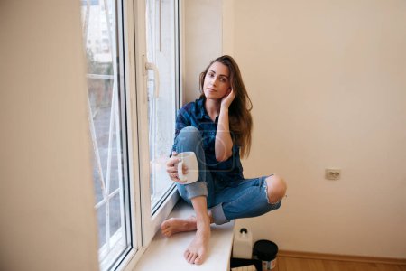 Téléchargez les photos : Jeune belle femme assise sur le rebord de la fenêtre, buvant et projetant de rénover la chambre. - en image libre de droit