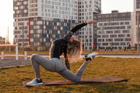 Photo for Young blonde woman is stretching on a playground on a yoga mat. Apartment buildings and people on background. Healthy energetic active lifestyle concept. - Royalty Free Image