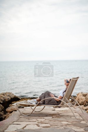 Foto de Feliz mujer relajada en gafas de sol y ropa de verano tumbado en la cama de sol entre las piedras cerca del mar ondulado. Viajar, personas y concepto de vacaciones. - Imagen libre de derechos