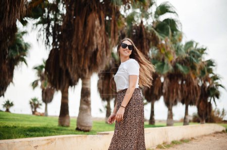 Photo for Portrait of positive young woman wearing summer clothes and sunglasses standing on hotel territory with palm trees around. Charming lady smiling and looking at camera. Winter at Cyprus - Royalty Free Image
