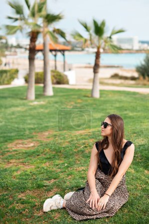 Foto de Encantadora mujer caucásica en gafas de sol y traje de verano sentado en la hierba cerca de la palmera en el territorio de lujo exótico resort. Concepto de verano, disfrute y relajación. - Imagen libre de derechos