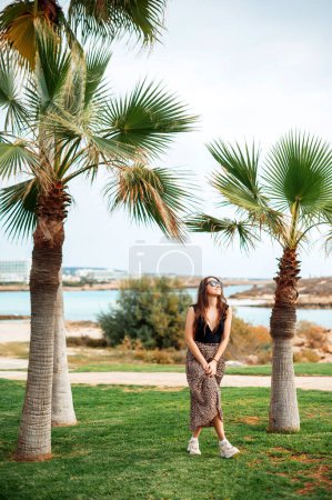 Photo for Full length portrait of beautiful caucasian woman in stylish clothes and sunglasses posing near palm tree on fancy hotel territory. Enjoyment from summer vacation. Cyprus winter time - Royalty Free Image