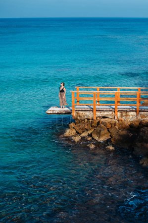 Foto de Caucasian young woman in sunglasses and summer dress standing on wooden pier near rough stones and waving sea. Amazing landscape on background. Cyprus winter time - Imagen libre de derechos