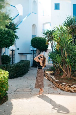 Téléchargez les photos : Adorable jeune femme en tenue d'été et lunettes de soleil posant sur le territoire de l'hôtel moderne avec des palmiers et de la verdure autour. Détente et plaisir pendant les vacances d'été. - en image libre de droit
