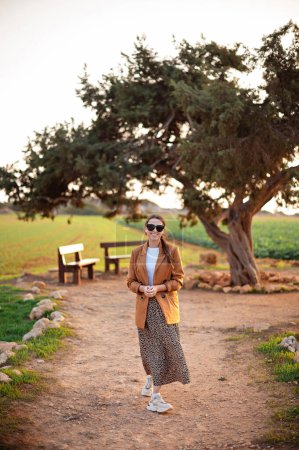 Foto de Mujer morena sonriente con ropa elegante y gafas de sol caminando cerca del árbol de los amantes en Ayia Napa. Turista femenina visitando lugar popular en Chipre. - Imagen libre de derechos