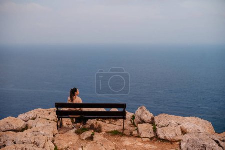 Foto de Vista trasera de la mujer morena sentada en un banco de madera y disfrutando de una hermosa vista al mar desde la roca. Increíble paisaje de verano durante las vacaciones. - Imagen libre de derechos