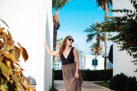 Photo for Pleasant dark haired woman wearing sunglasses and stylish dress standing among palm trees on hotel territory. Luxury resort for summer vacation. - Royalty Free Image