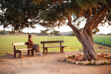 Foto de Hermosa mujer joven en gafas de sol y ropa elegante sentado en el banco de madera cerca del viejo árbol de los amantes en Chipre. Viajar, personas y concepto de vacaciones. - Imagen libre de derechos