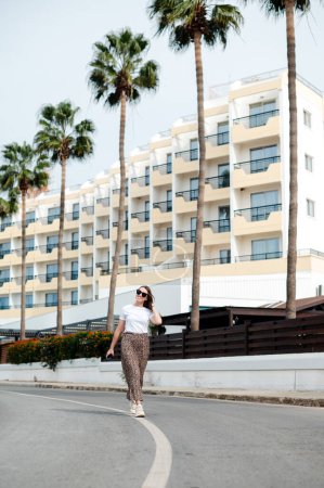 Foto de Mujer joven sonriente en gafas de sol y ropa elegante caminando por el territorio del hotel moderno con palmeras alrededor. Vacaciones de verano en un resort tropical. - Imagen libre de derechos