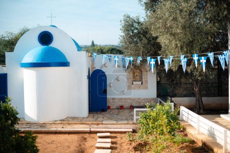 Foto de Iglesia chipriota tradicional con techo azul y pared blanca. Banderas simbólicas colgando cerca con olivos creciendo alrededor. - Imagen libre de derechos