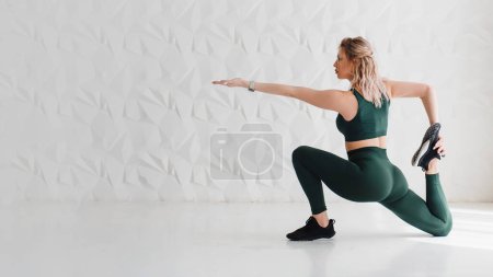 Photo for Beautiful sporty woman in crop top and green leggings doing stretching for legs while sitting on white floor. Concept of stretching workout and healthy lifestyle - Royalty Free Image